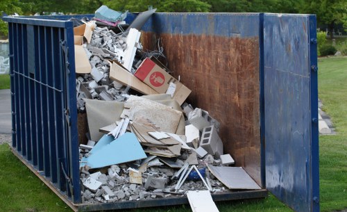 A large, open industrial waste container situated outdoors on a grassy area, filled with a scattered mixture of construction debris and household rubbish including broken bricks, plasterboard, pieces of wood, cardboard boxes, plastic containers, and various discarded materials. The container has a blue metal frame with rusted interior surfaces, and its height exceeds that of nearby bushes and trees, suggesting a significant capacity for rubbish removal services. The scene is set on a clear day with natural lighting, capturing the environmental context of waste disposal, typical of commercial waste removal operations in Knightsbridge and surrounding London areas. The detailed textures of the debris range from smooth cardboard to rough concrete and the container’s weathered finish emphasizes its industrial use, aligning with professional rubbish collection and waste management services provided by Commercial Waste Removal Knightsbridge.