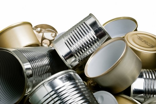 The image displays a close-up of multiple metal cans, some with ridged sides and others smooth, stacked and scattered on a surface. The cans vary in size and are predominantly silver and gold in colour, with some lids visible on a few of them. The background appears neutral and plain, emphasizing the metallic textures and finishes of the cans. The scene suggests an industrial or waste collection context, relevant to rubbish removal services, with the cans arranged in a slightly disorganized manner. The metallic sheen and reflective surfaces highlight the material composition, likely aluminium or tin, commonly found in household or commercial waste. The setting appears to be in a warehouse or collection point, typical in waste removal operations in Knightsbridge or nearby areas. Commercial Waste Removal Knightsbridge offers services that manage such metallic waste efficiently, fitting into a typical urban environment for rubbish disposal or recycling activities. The lighting is even, with no harsh shadows, allowing clear detail of the cans' textures and finishes to be seen clearly.
